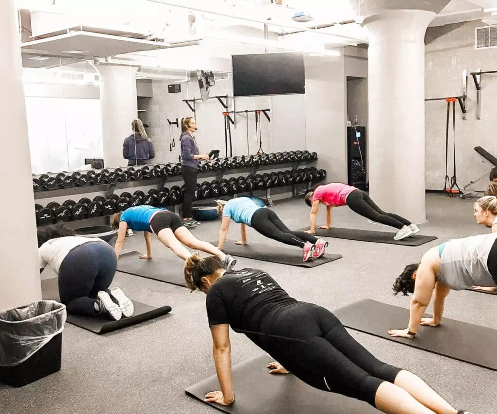 7 people participating in a fitness class with their hands and feet on mats with an instructor with a microphone at the front of the room. Dumbbells lining the mirrored wall 