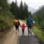 two adults holding the hands of a kid walking down a path in the mountains on a rainy day